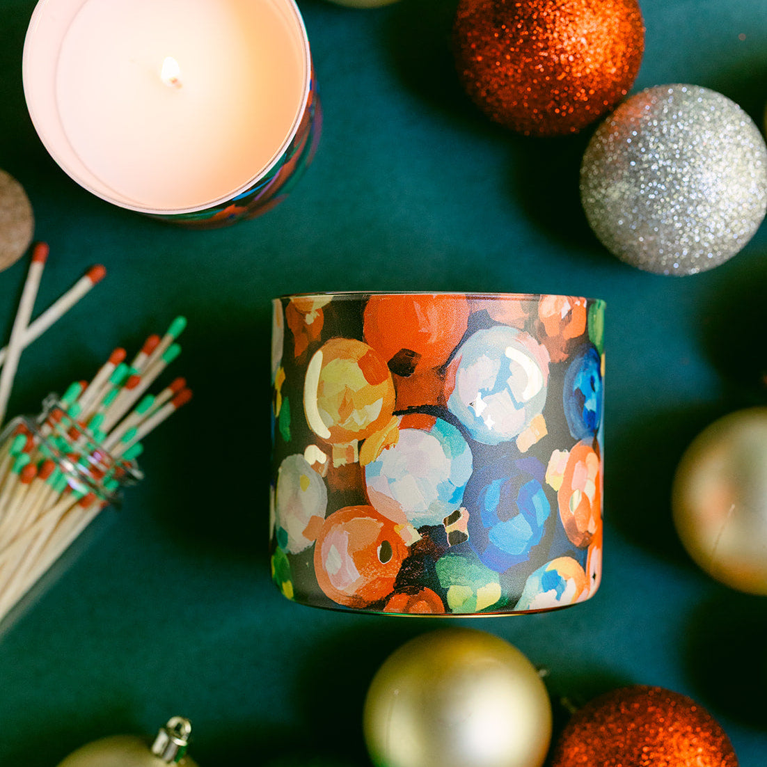 Decorative candle with colorful design surrounded by Christmas ornaments on a green surface
