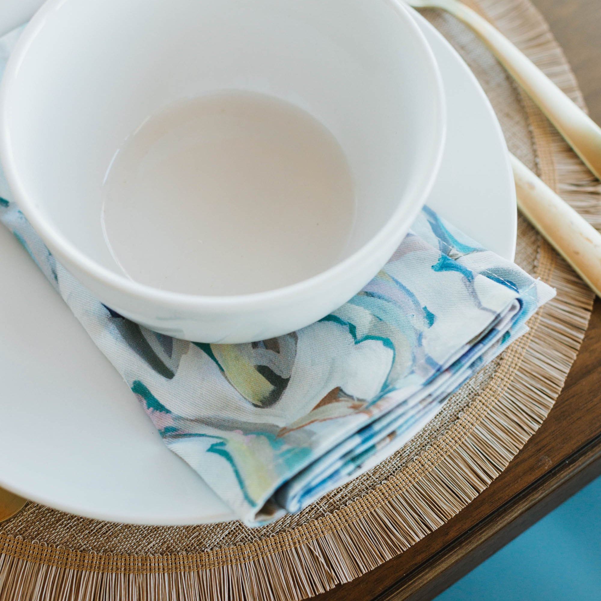 White bowl on a decorative plate with a fish-pattern napkin and spoon on a wooden table.