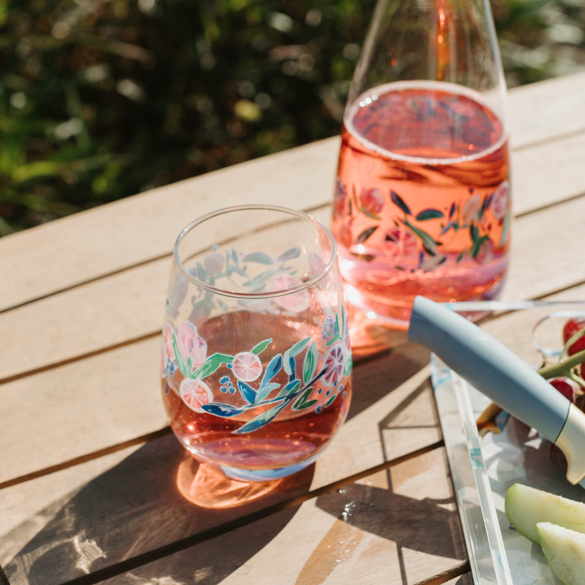 Stemless wine glass and carafe with floral design on a wooden table outdoors.
