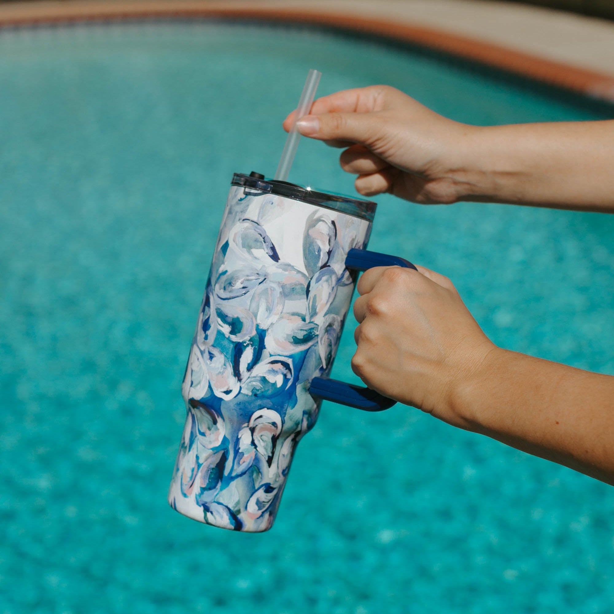 Person holding a Kim Hovell blue shell-patterned tumbler with lid and straw by a pool