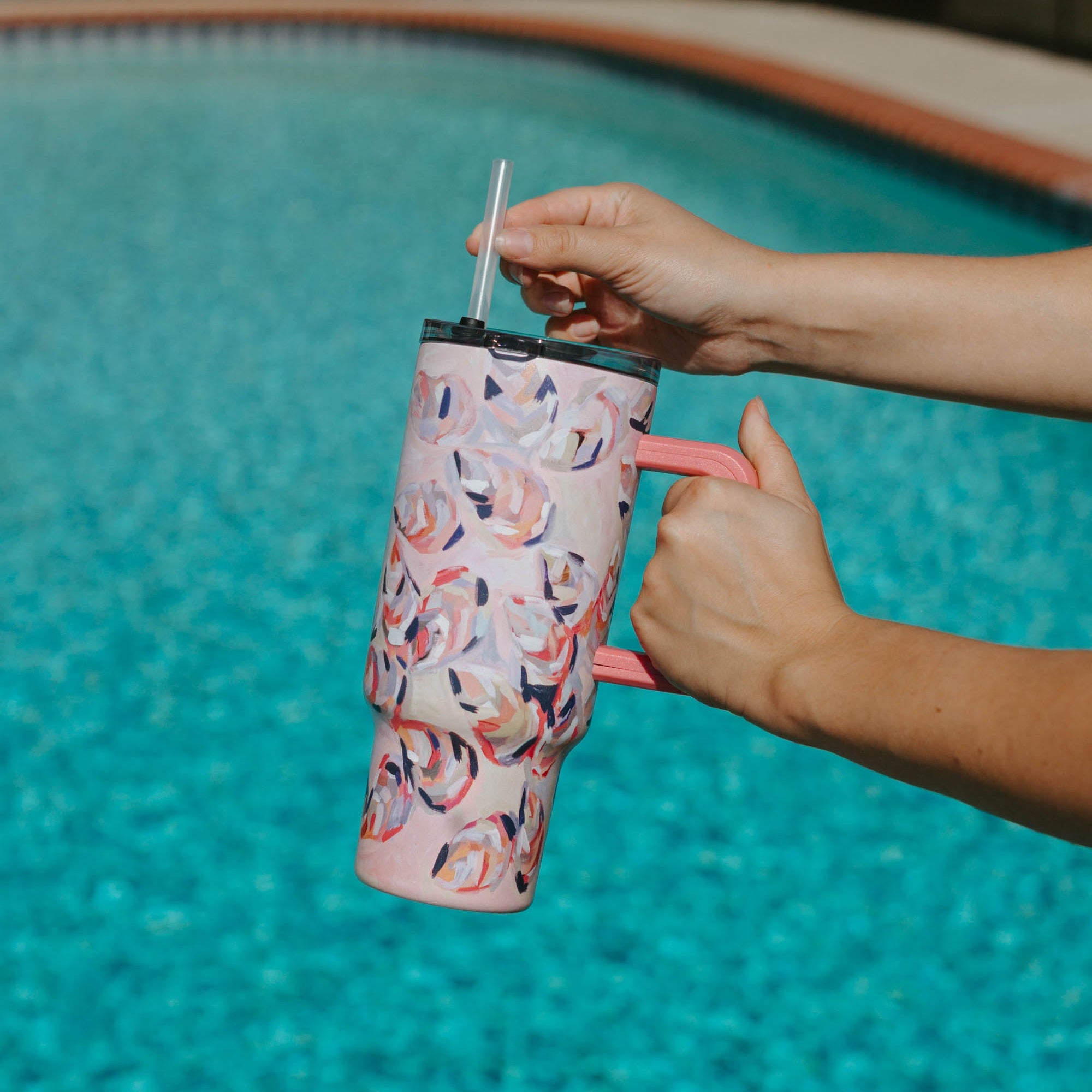 Person holding a Kim Hovell Peach Gems pink shell-patterned tumbler with lid and straw by a pool