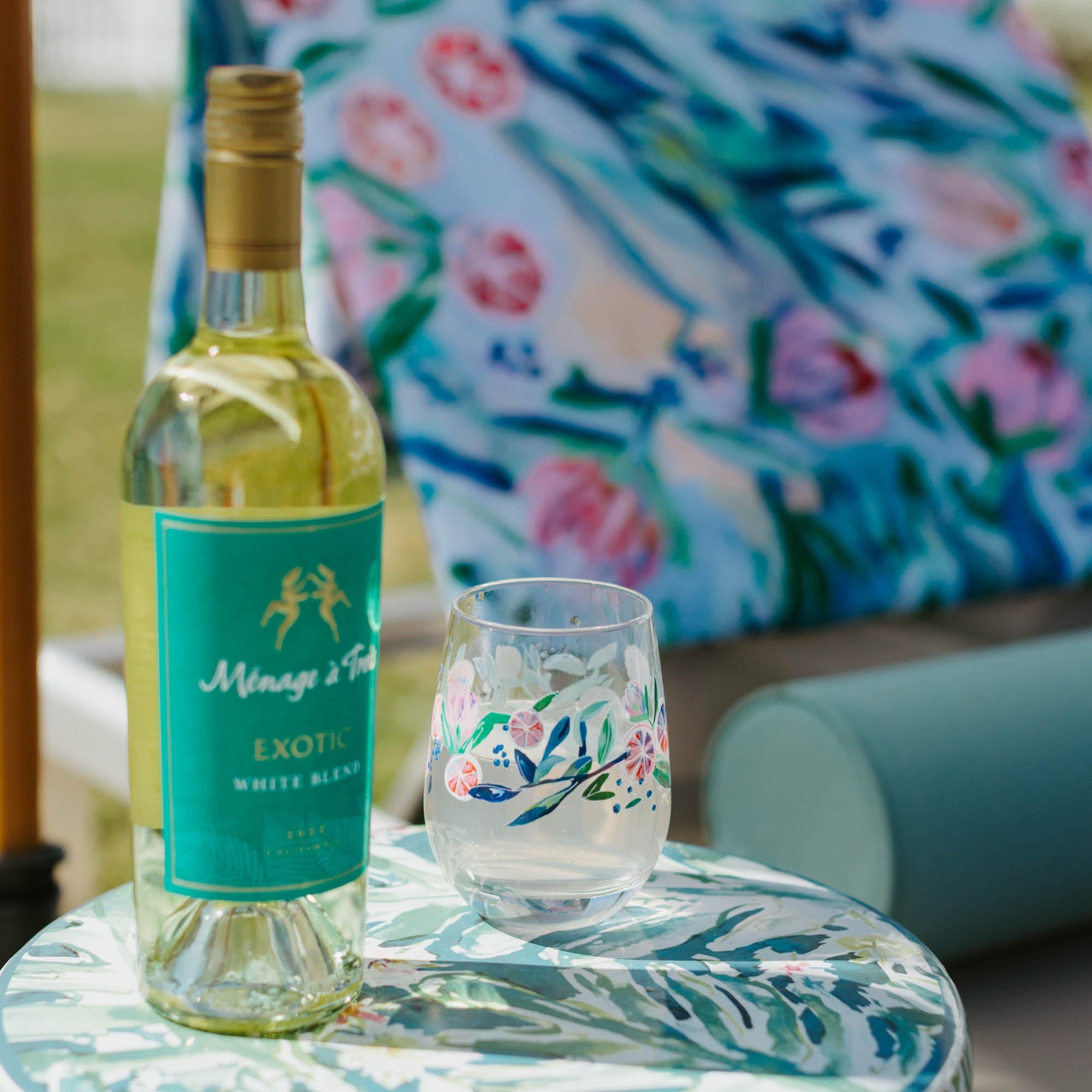 Wine bottle and glass on a floral table with a colorful chair in the background