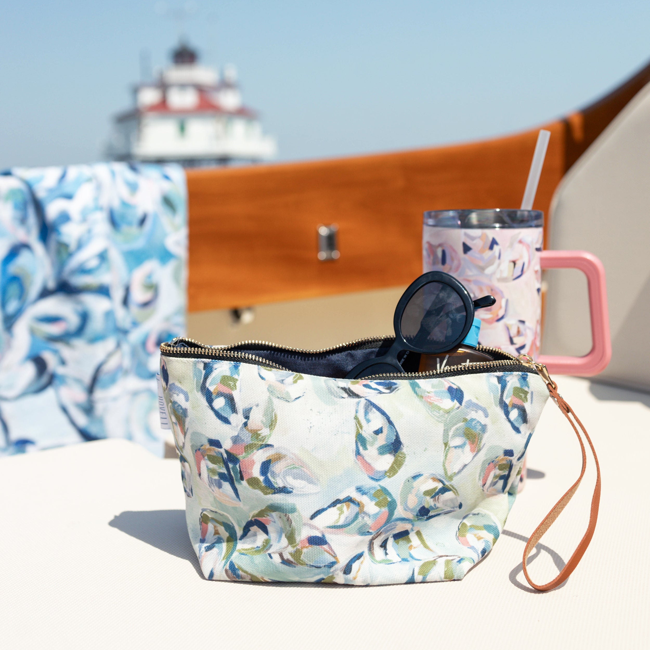Kim Hovell Gulf Cluster patterned makeup bag with sunglasses and a pink tumbler on a boat with a lighthouse in the background