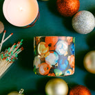 Decorative candle with colorful design surrounded by Christmas ornaments on a green surface