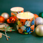Decorative candles with colorful designs surrounded by Christmas ornaments on a green surface.