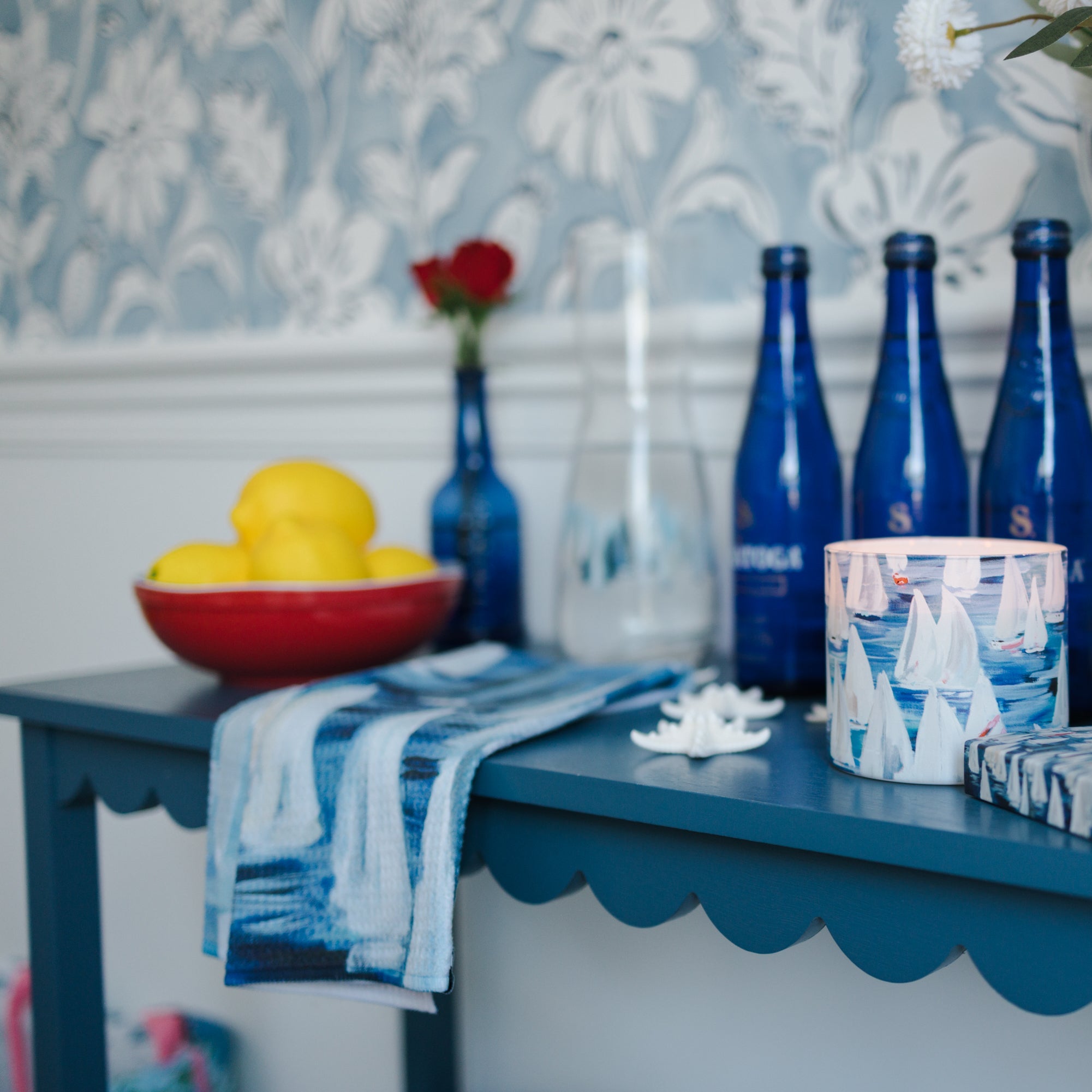 Decorative setting with blue bottles, a white pitcher, and a bowl of fruit on a shelf.