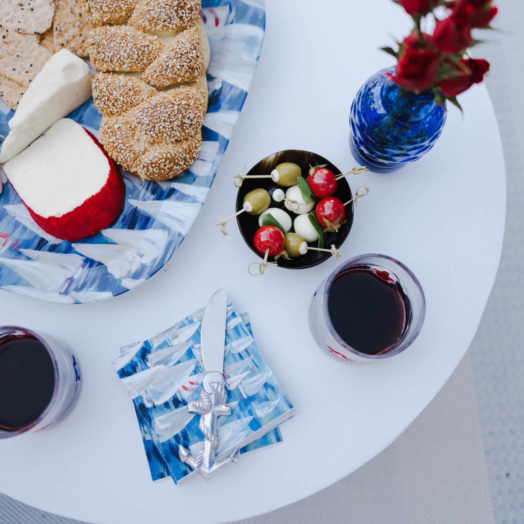 Round white table with a plate of food, a vase of flowers, and glasses of red wine.