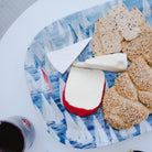 Platter with assorted cheeses, crackers, and a glass of red wine on a white table.