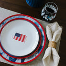 Stack of plates with American flag design on a table setting with a napkin and glass.