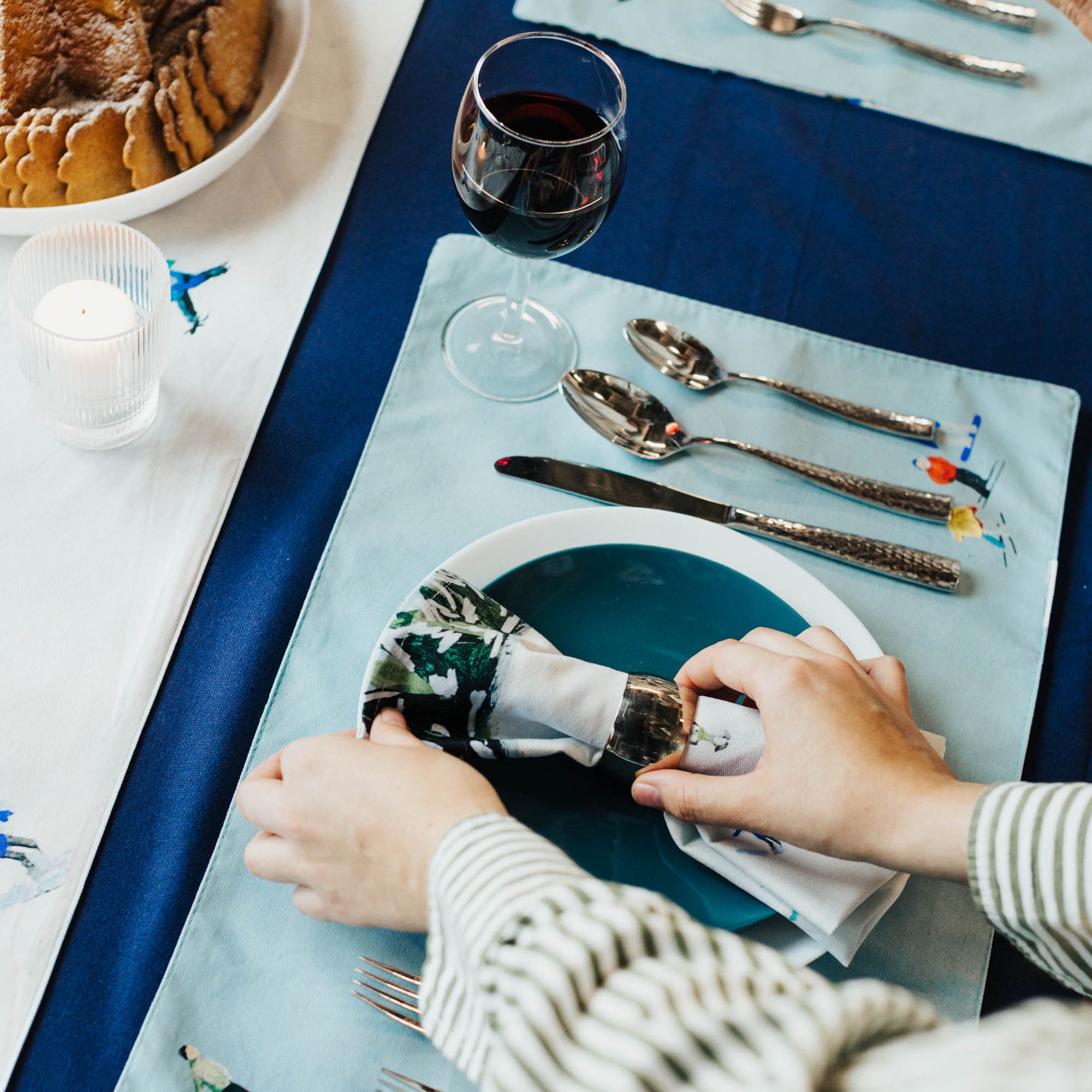 Person setting a table with cutlery on a blue tablecloth and decorative placemat