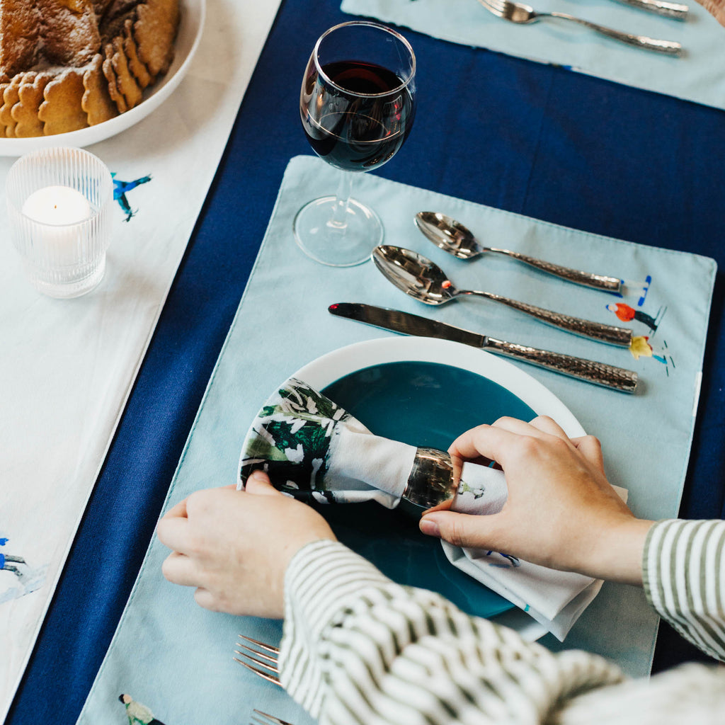 Person setting a table with cutlery on a blue tablecloth and decorative placemat