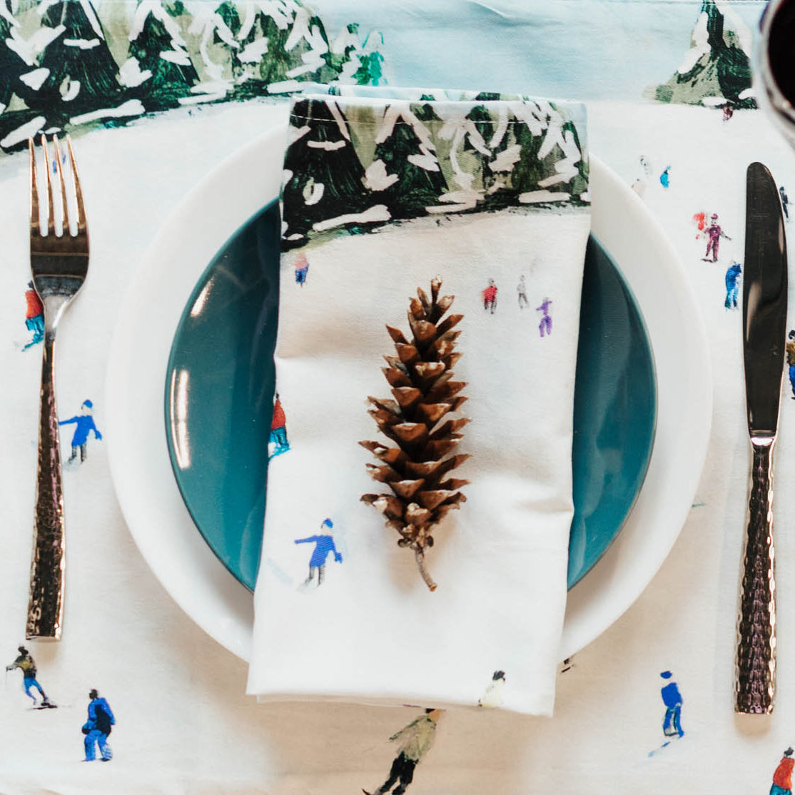 Table setting with a pine cone on a white napkin featuring skiing figures, surrounded by decorative elements.