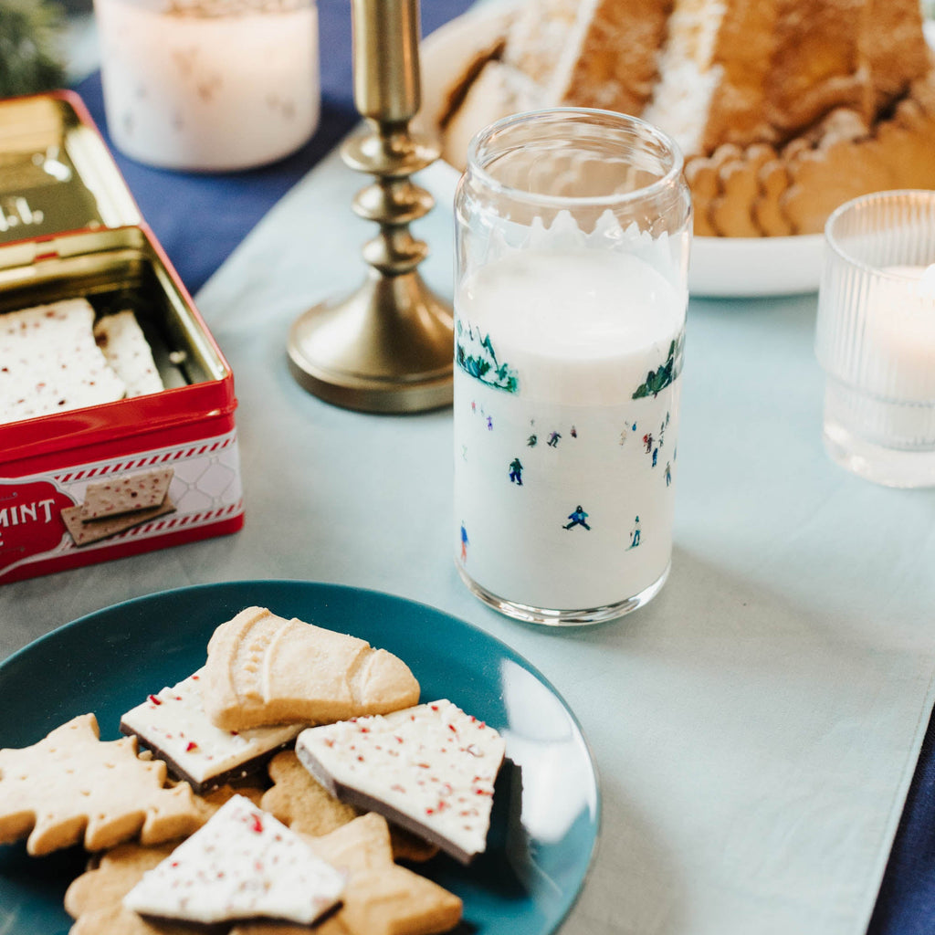 Plate of cookies with a glass of milk on a table setting with candles and a cake.