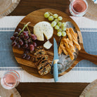 Wooden charcuterie board with cheese, grapes, and crackers on a table with a blue and white checkered tablecloth.