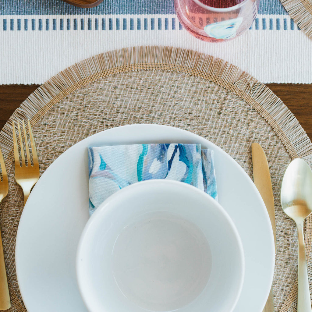 Dining table setting with white plates, gold cutlery, fish-pattern napkin, and a woven placemat.