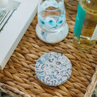 Decorative glass coaster with blue floral design on a woven tray with a book and bottle.