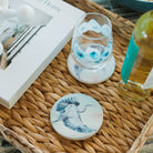 Decorative coasters with bird designs on a woven tray with a book and bottle in the background.