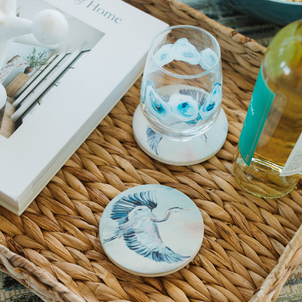 Decorative coasters with bird designs on a woven tray with a book and bottle in the background.