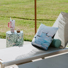 Outdoor lounge chairs with sailboat pillow and a large umbrella on a patio, with a scenic background.