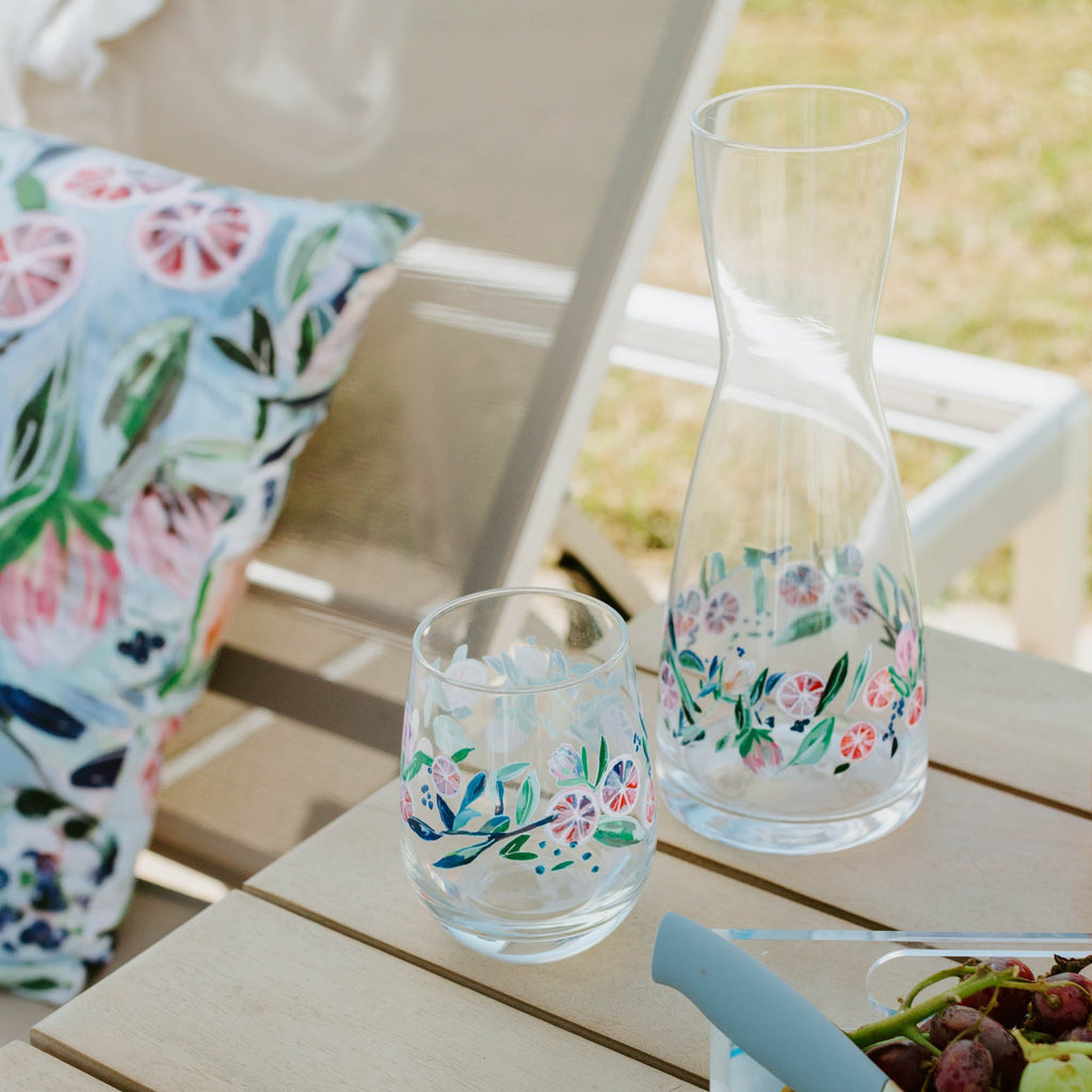 Clear glass carafe and stemless wine glasses with floral designs on a wooden table.