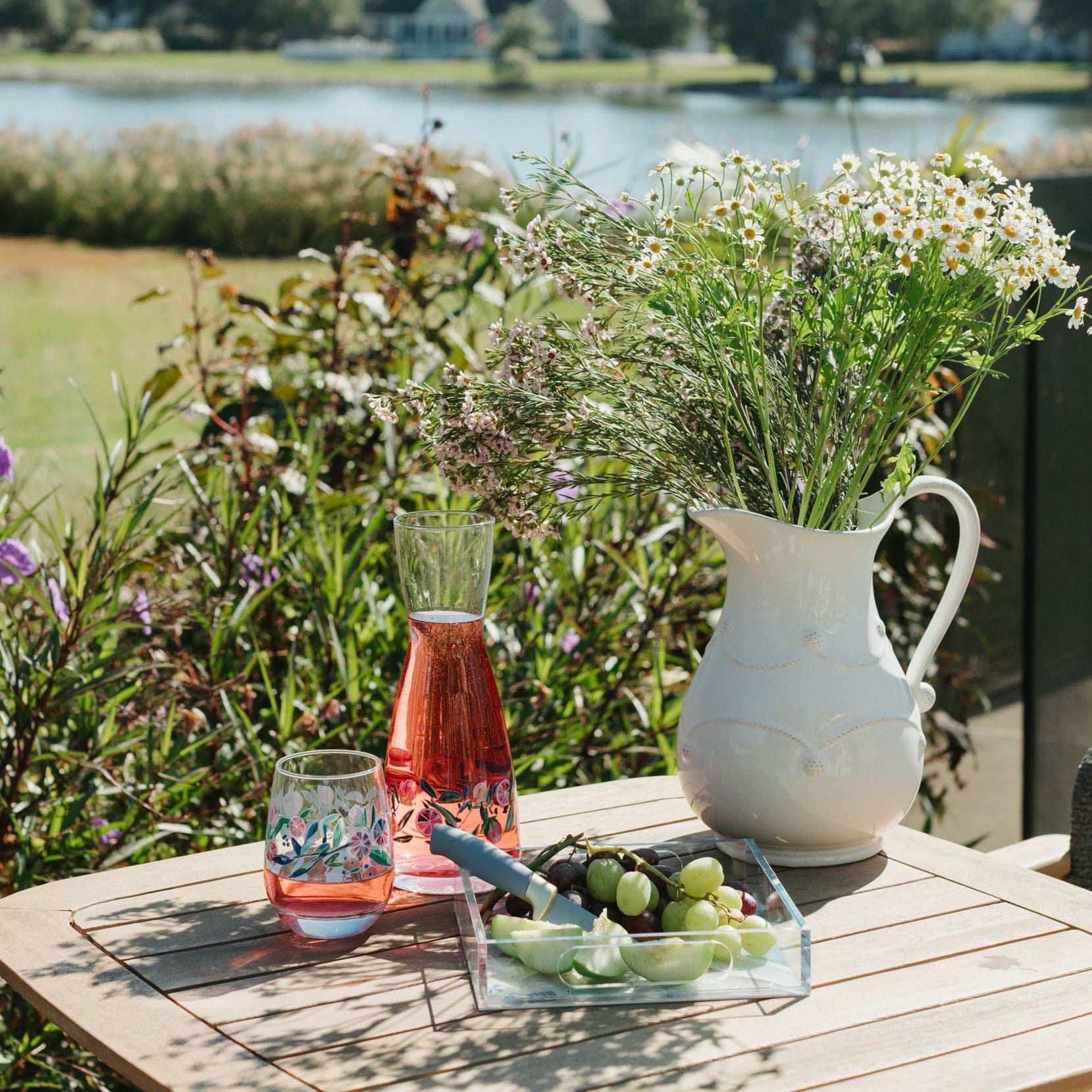 Outdoor setting with a wooden table by a lake, featuring a pitcher, glasses, and fruit.