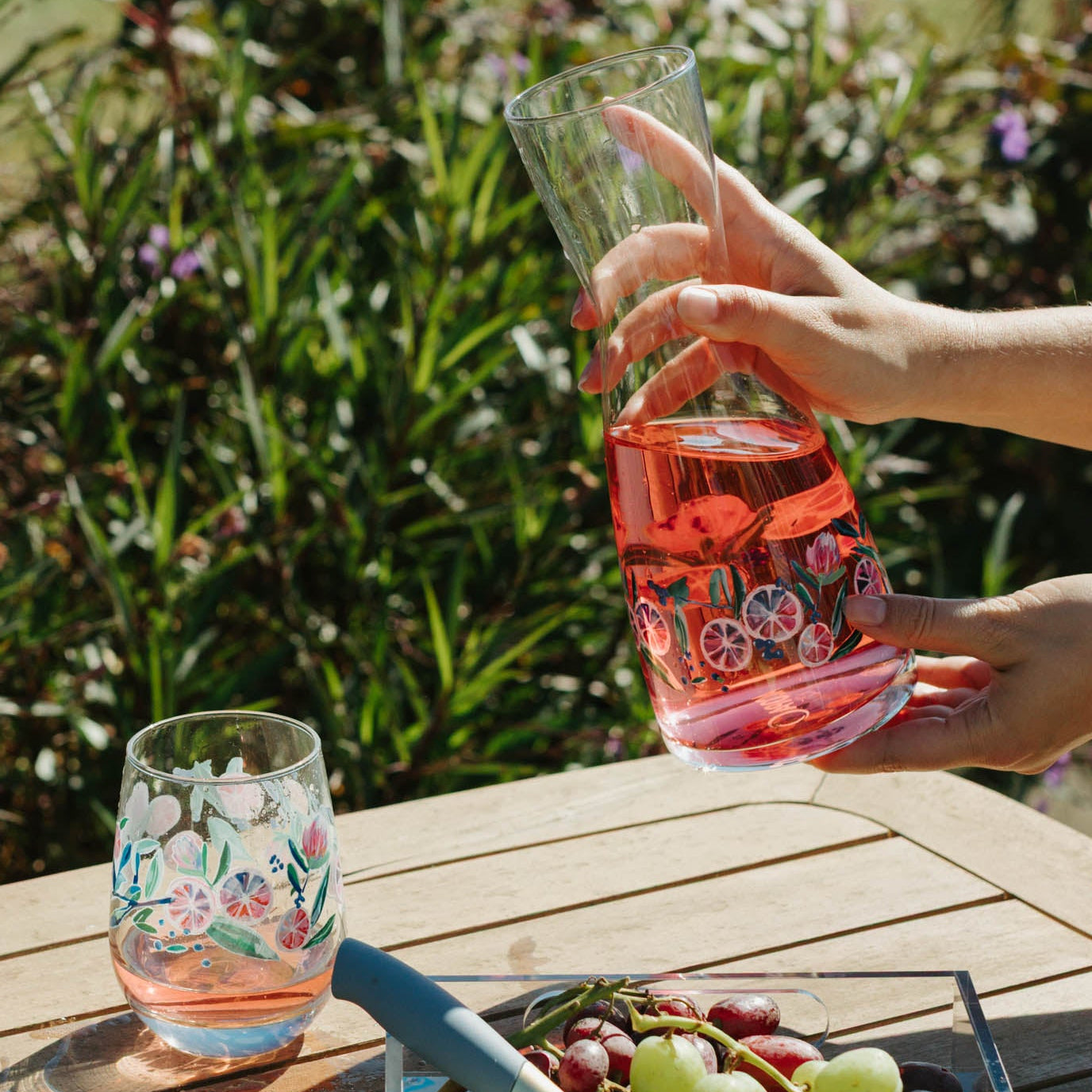 Person pouring pink drink into a glass on a wooden table with a scenic background