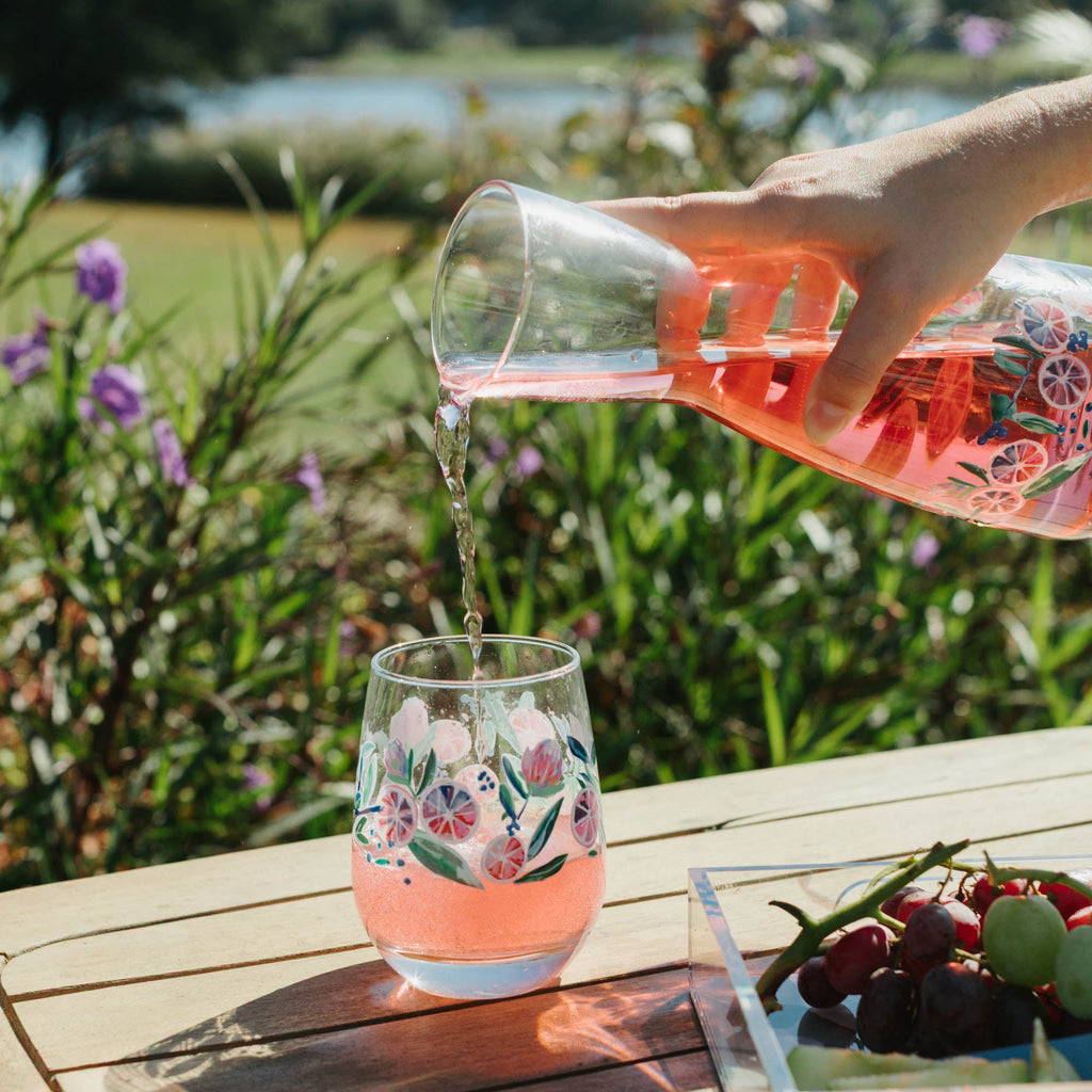 Hand pouring pink floral-patterned drink into a glass on a wooden table with a scenic background.