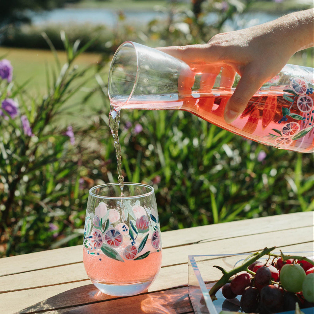 Hand pouring pink floral-patterned drink into a glass on a wooden table with a scenic background.