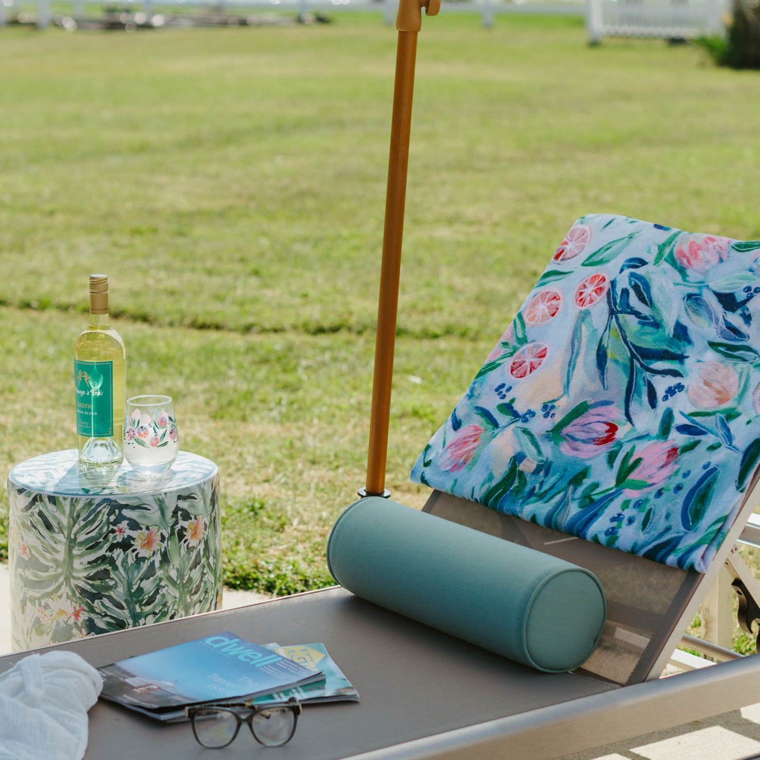 Lounge chair with a floral towel, bottle, and sunglasses under a white umbrella by a lake.