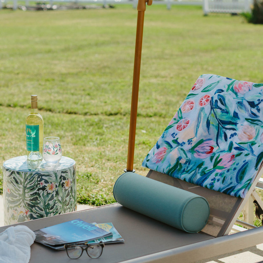 Lounge chair with a floral towel, bottle, and sunglasses under a white umbrella by a lake.