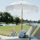 Outdoor lounge area with white umbrella, chairs, and decorative pillows by a pond.