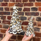 Two hands holding up decorative art made of many white and gray oyster shells, shown against a rustic red-brown brick wall.